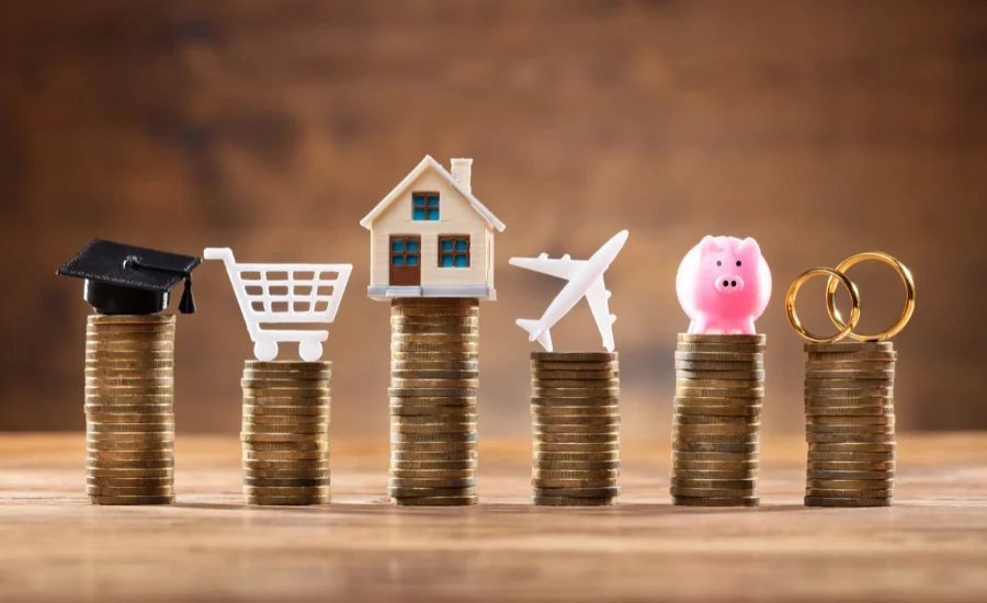Pilhas de moedas com ícones de elementos de viagem no topo: um chapéu de formatura, um carrinho de compras, uma casa, um avião e um cofrinho, simbolizando os custos e a proteção oferecida pelo seguro viagem.