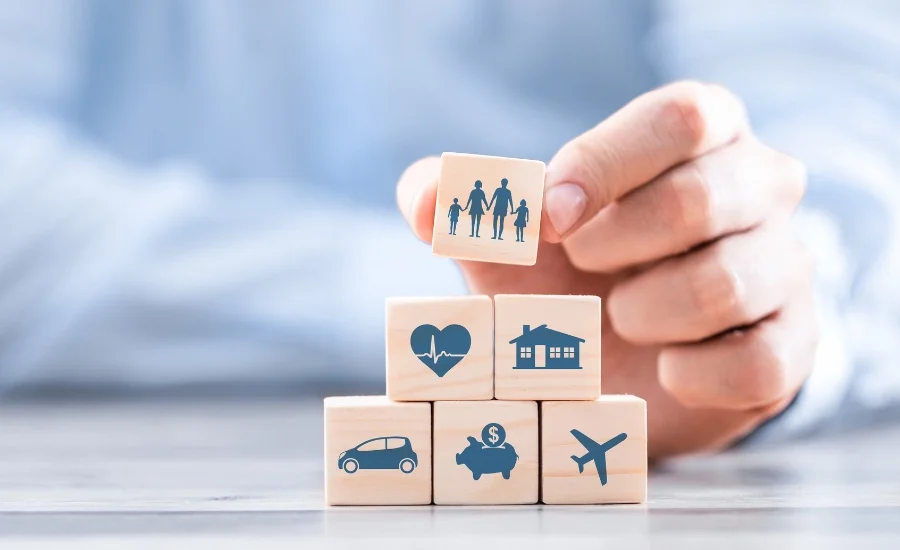Mão colocando um bloco de madeira ilustrado com figuras humanas sobre outros blocos com ícones de saúde, casa, carro, economia e avião.
